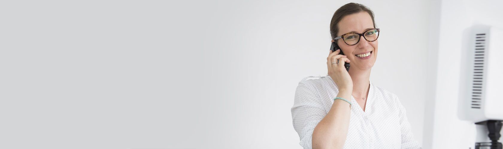 A woman in glasses smiling while talking on the phone in a bright pharmacy setting.