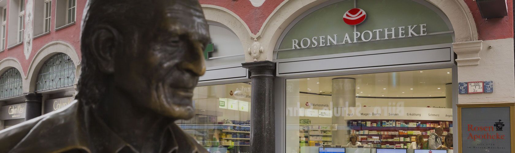Bronzestatue vor der Rosen Apotheke in einer belebten Straße. Die Apotheke ist modern gestaltet und gut besucht.