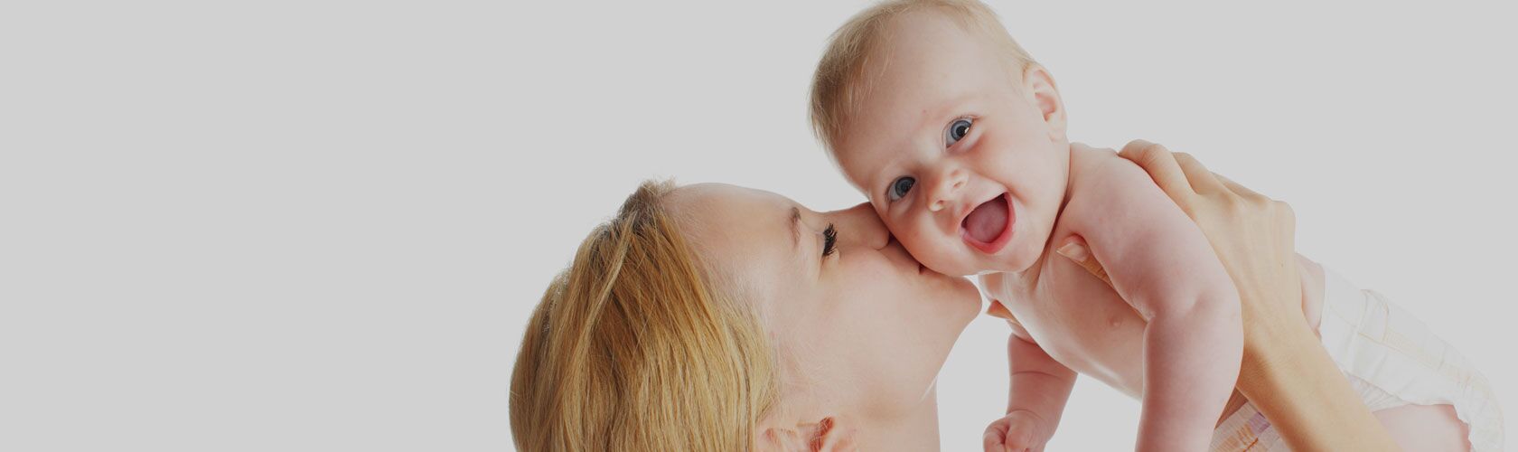 Mutter hält lächelndes Baby in den Armen und gibt ihm einen Kuss auf die Wange vor weißem Hintergrund.