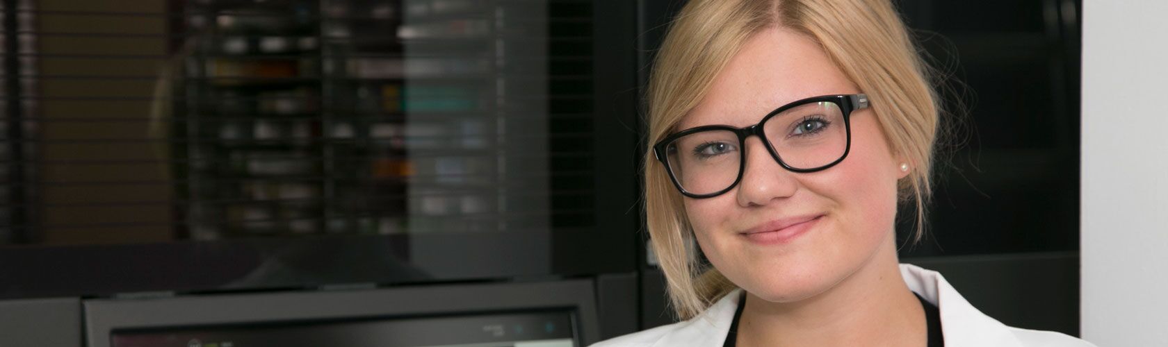 Junge Frau mit Brille lächelt in die Kamera, im Hintergrund moderne Büroumgebung, symbolisiert beruflichen Erfolg.